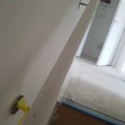 Świeżo zamontowany, biały karnisz na ścianie w trakcie prac wykończeniowych, zabezpieczony ż&oacute;łtą taśmą malarską, widoczne ślady po remoncie na podłodze i w tle otwarte drzwi do kolejnego...