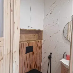 Nowoczesna łazienka z czarną toaletą wiszącą, bidetką w kolorze czarnym, białymi płytkami imitującymi marmur ze złotymi żyłami, drewnianą zabudową, białymi szafkami i okrągłym lustrem.