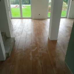 Ułożona podłoga z płytek imitujących drewno w jasnym, nowym wnętrzu z dużymi oknami wychodzącymi na zieleń. Widoczne ślady po czyszczeniu na powierzchni płytek.