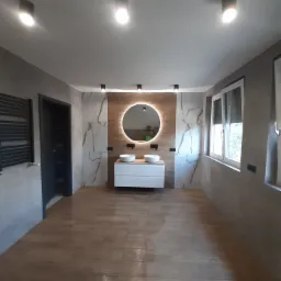 Nowoczesna łazienka z podświetlanym okrągłym lustrem, dwoma białymi umywalkami nablatowymi, białą szafką pod umywalką, płytkami imitującymi marmur na ścianach i drewnopodobnymi płytkami na podłodze...