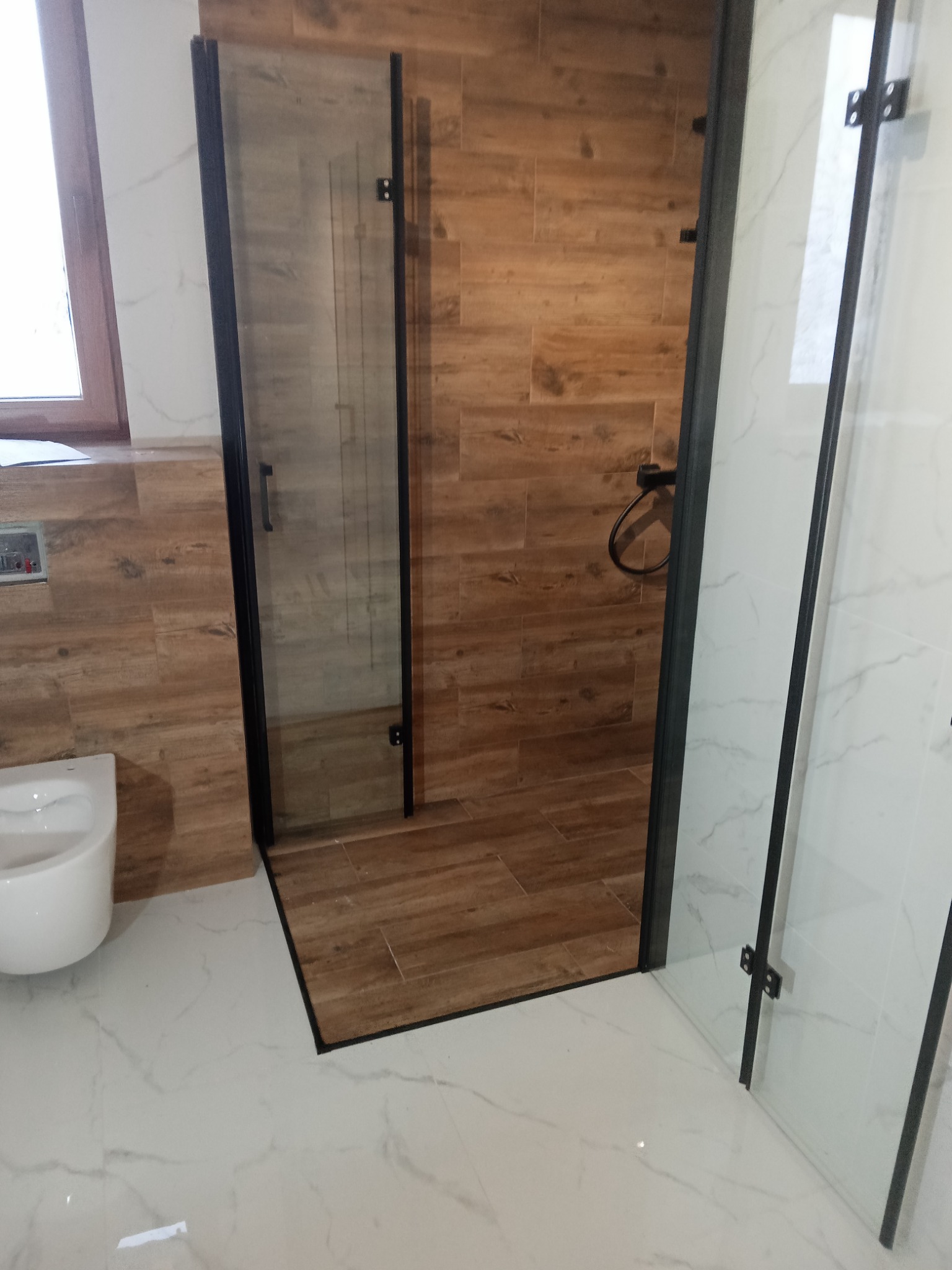 Nowoczesna kabina prysznicowa z czarnymi profilami i drewnopodobnymi płytkami wewnątrz, kontrastująca z białymi marmurkowymi płytkami na zewnątrz kabiny i na podłodze, z widoczną toaletą w tle.