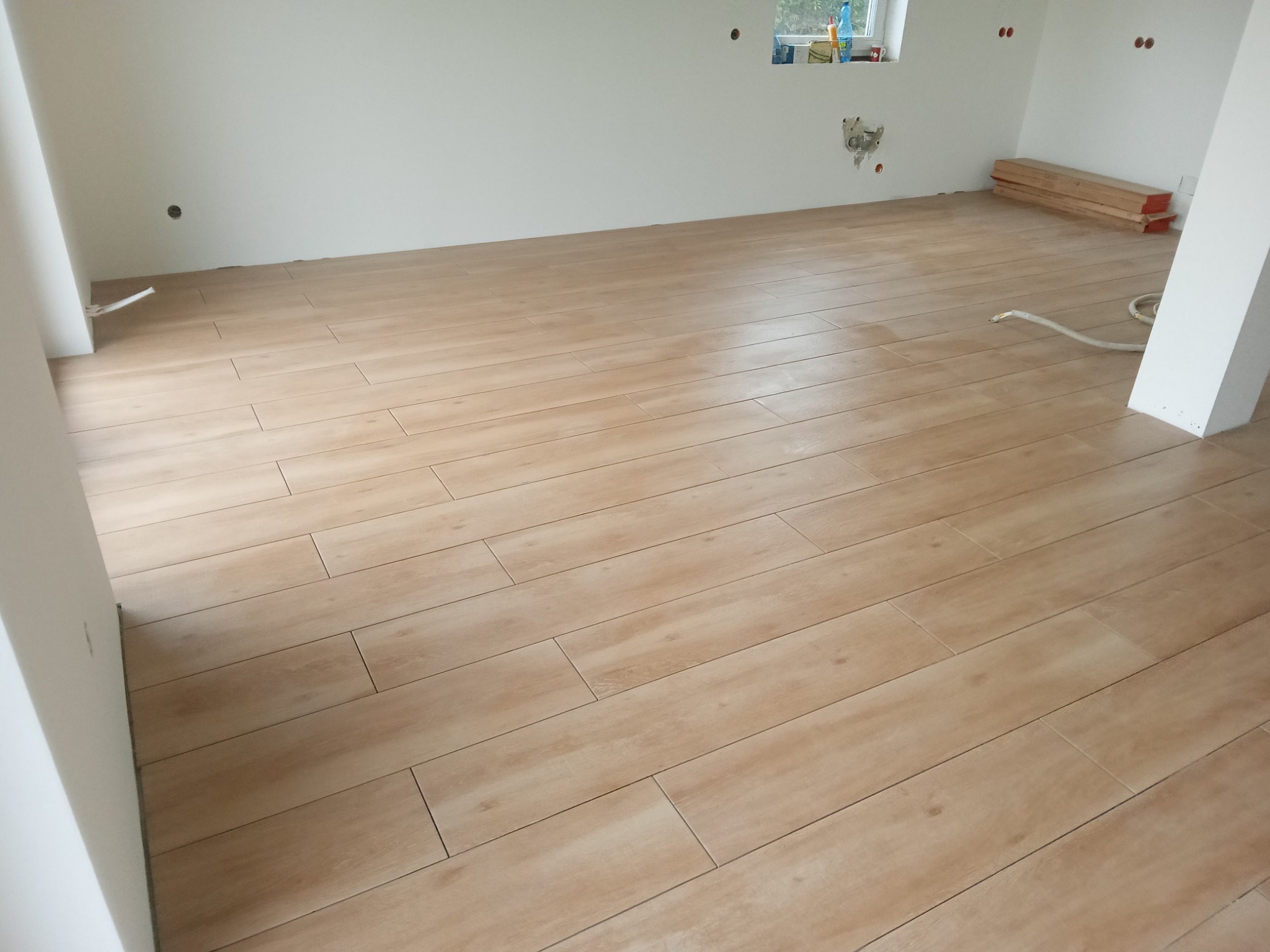 Ułożona podłoga z płytek imitujących drewno w jasnym odcieniu, widoczne przygotowane panele podłogowe przy ścianie, surowe ściany z wyprowadzonymi instalacjami.