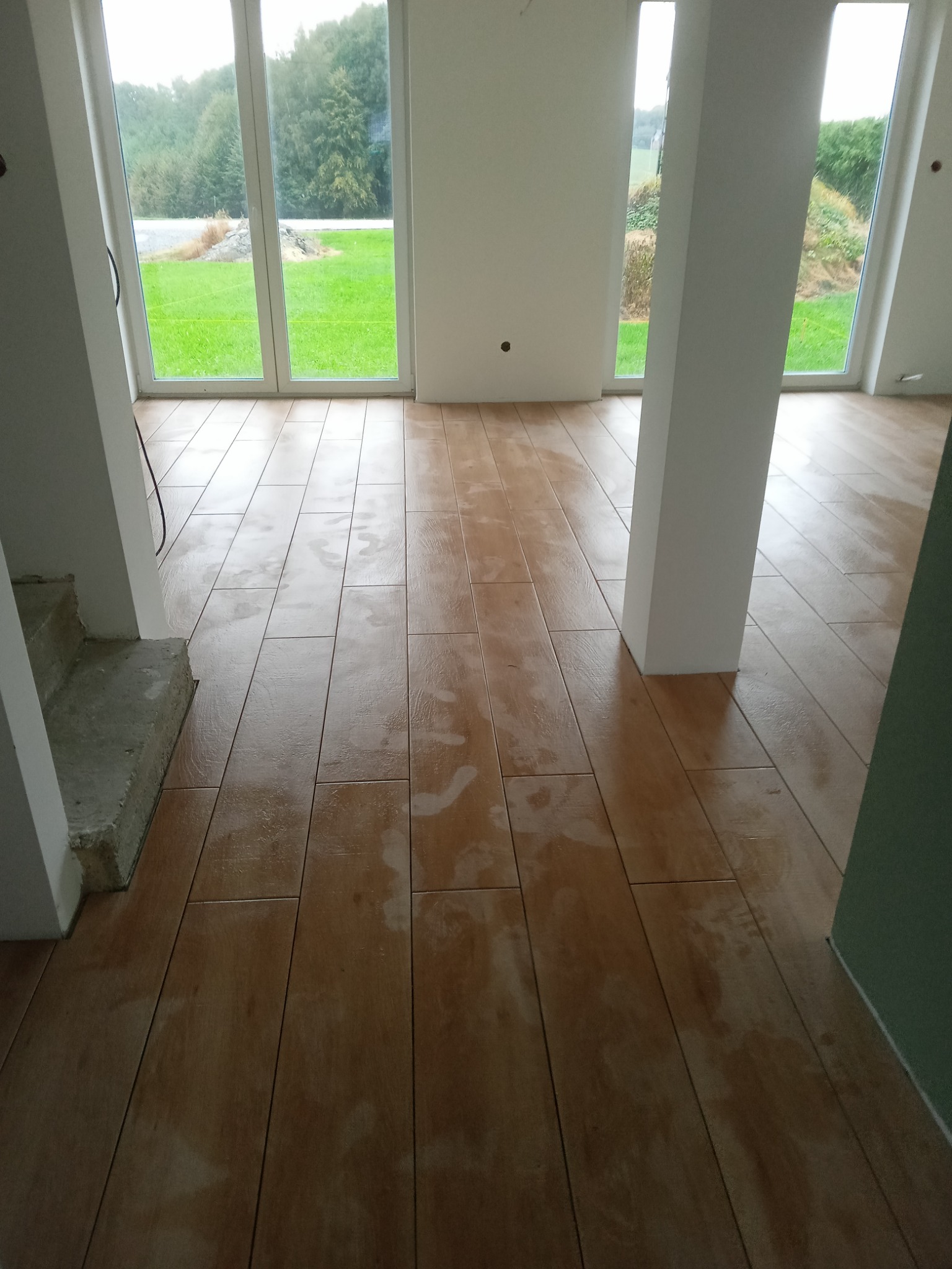 Ułożona podłoga z płytek imitujących drewno w jasnym, nowym wnętrzu z dużymi oknami wychodzącymi na zieleń. Widoczne ślady po czyszczeniu na powierzchni płytek.