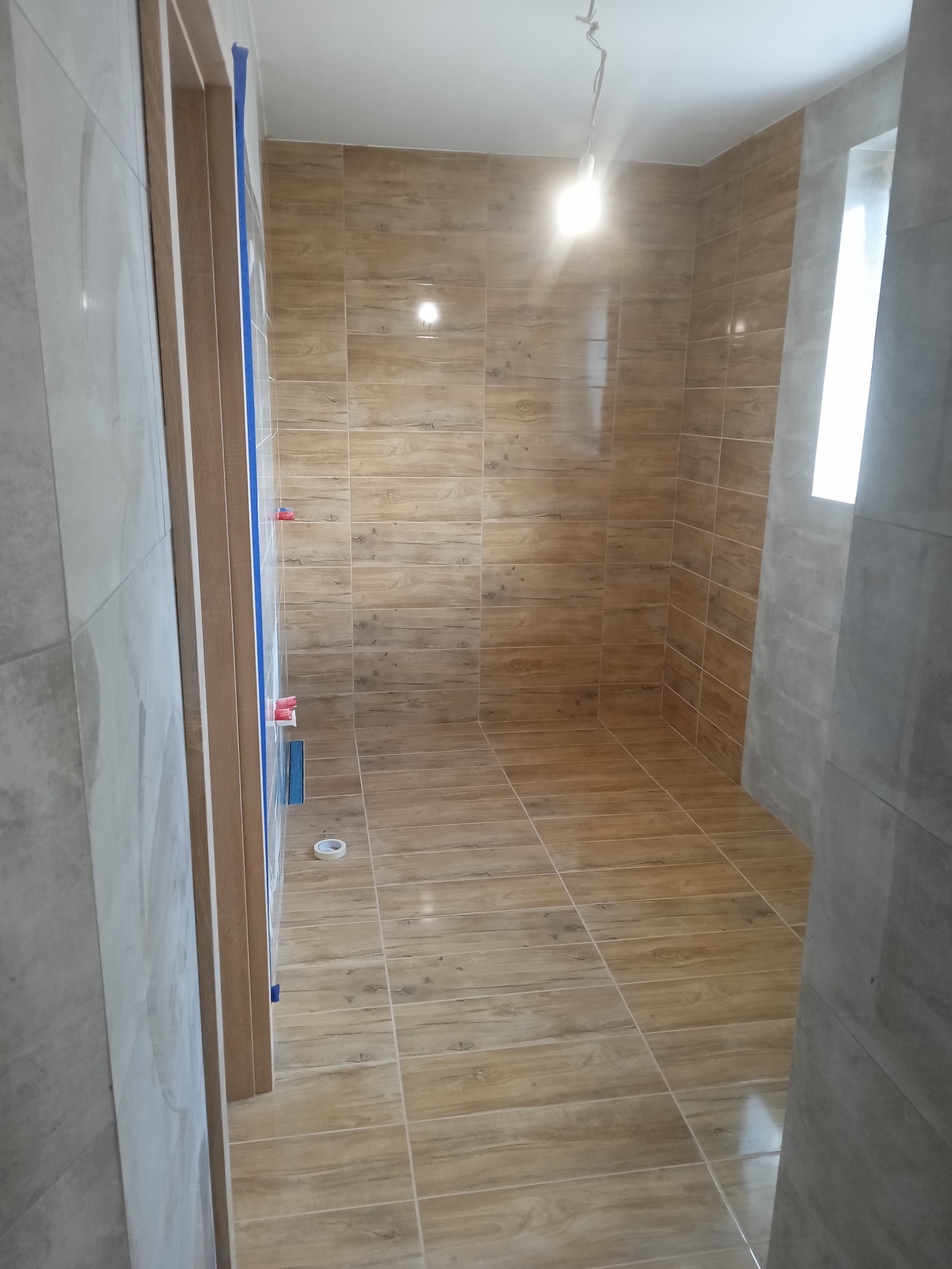 Wnętrze łazienki w trakcie prac wykończeniowych z brązowymi płytkami imitującymi drewno na ścianach i podłodze, widoczna taśma malarska zabezpieczająca ościeżnicę drzwi, surowa żarówka zwisająca...