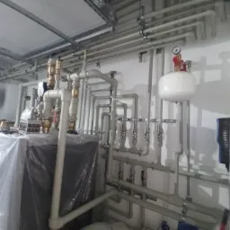 Instalacja grzewcza w kotłowni: białe rury, zawory, manometr, zbiornik wyrównawczy i piec owinięty folią ochronną.