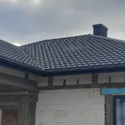 Nowy, grafitowy dach z blachodachówki modułowej i komin wentylacyjny na surowym budynku z betonu komórkowego, widoczne przygotowania do ocieplenia.