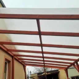 Zadaszenie tarasu z brązowych drewnianych belek i przezroczystego poliwęglanu, widok od dołu na tle budynku.