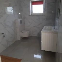 Wykończona łazienka z marmurowymi płytkami ściennymi, podwieszanym WC, szafką z umywalką i grzejnikiem drabinkowym. Fragment drewnianej podłogi widoczny w rogu.