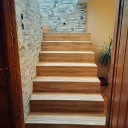 Schody wykończone panelami imitującymi drewno, z dekoracyjną ścianą z kamienia w odcieniach szarości i beżu, widok z dołu przez otwarte drewniane drzwi.