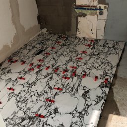 Rem-Compleks Piotr Sojka - Układanie płytek podłogowych imitujących marmur z użyciem czerwonych klinów poziomujących w trakcie prac w remontowanej łazience z odsłoniętymi instalacjami.