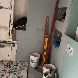 Rem-Compleks Piotr Sojka - Fragment łazienki w trakcie prac: ułożone płytki imitujące marmur z czerwonymi krzyżykami dystansowymi, ściana częściowo pokryta szarą gładzią, widoczne poziomice, puszki z materiałami budowlanymi...