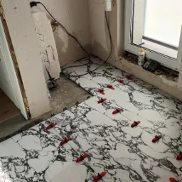 Układanie białych płytek podłogowych imitujących marmur, z czerwonymi krzyżykami dystansowymi, widoczne niedokończone ściany i instalacje elektryczne.