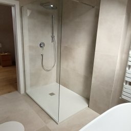 Rem-Compleks Piotr Sojka - Beżowa łazienka po remoncie: kabina prysznicowa typu walk-in z deszczownicą, biała wanna i toaleta. Widoczna żarówka na kablu. Minimalistyczny design.