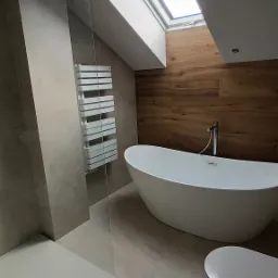 Nowoczesna łazienka na poddaszu z wolnostojącą wanną, kabiną prysznicową i drewnianą okładziną ściany. Widoczne okno dachowe i grzejnik drabinkowy.