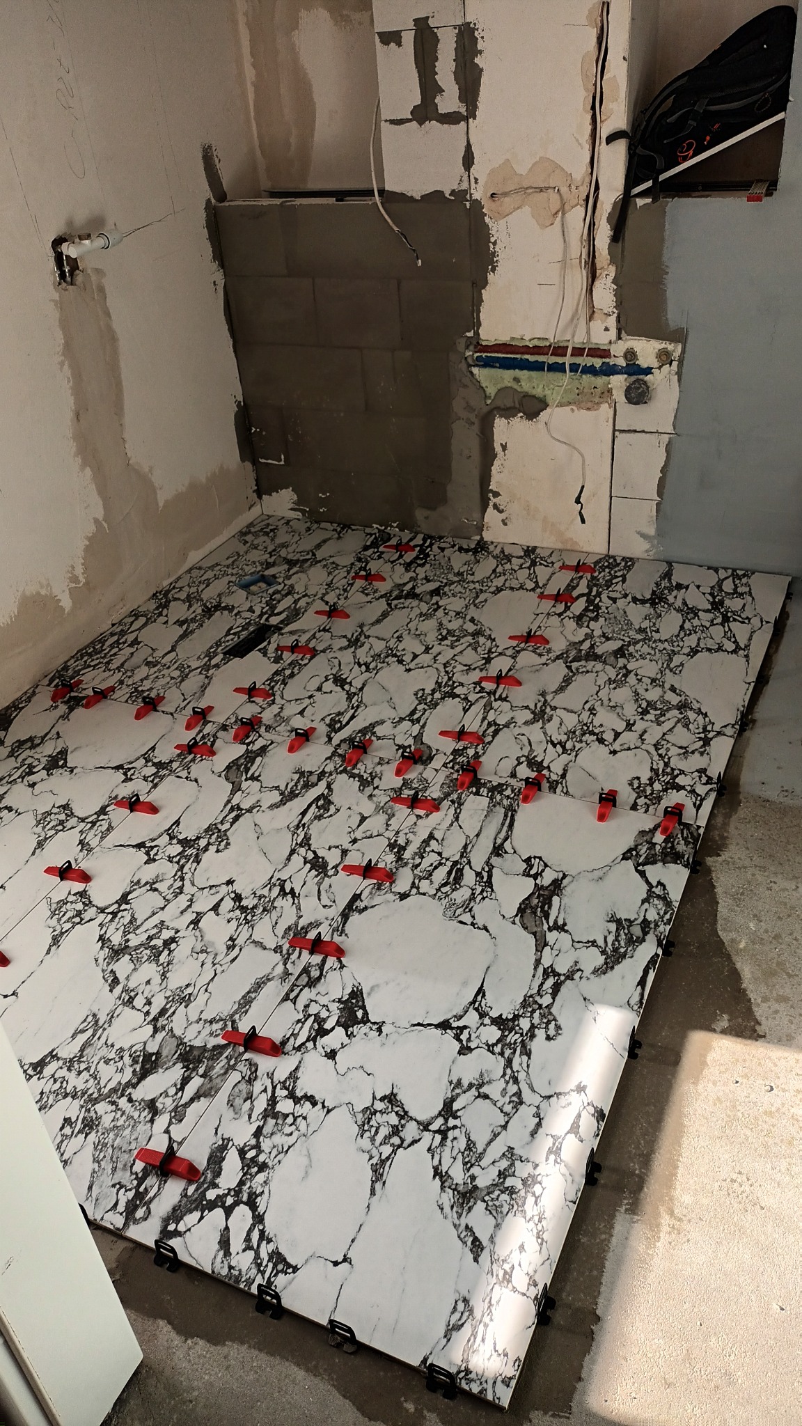 Układanie płytek podłogowych imitujących marmur z użyciem czerwonych klinów poziomujących w trakcie prac w remontowanej łazience z odsłoniętymi instalacjami.