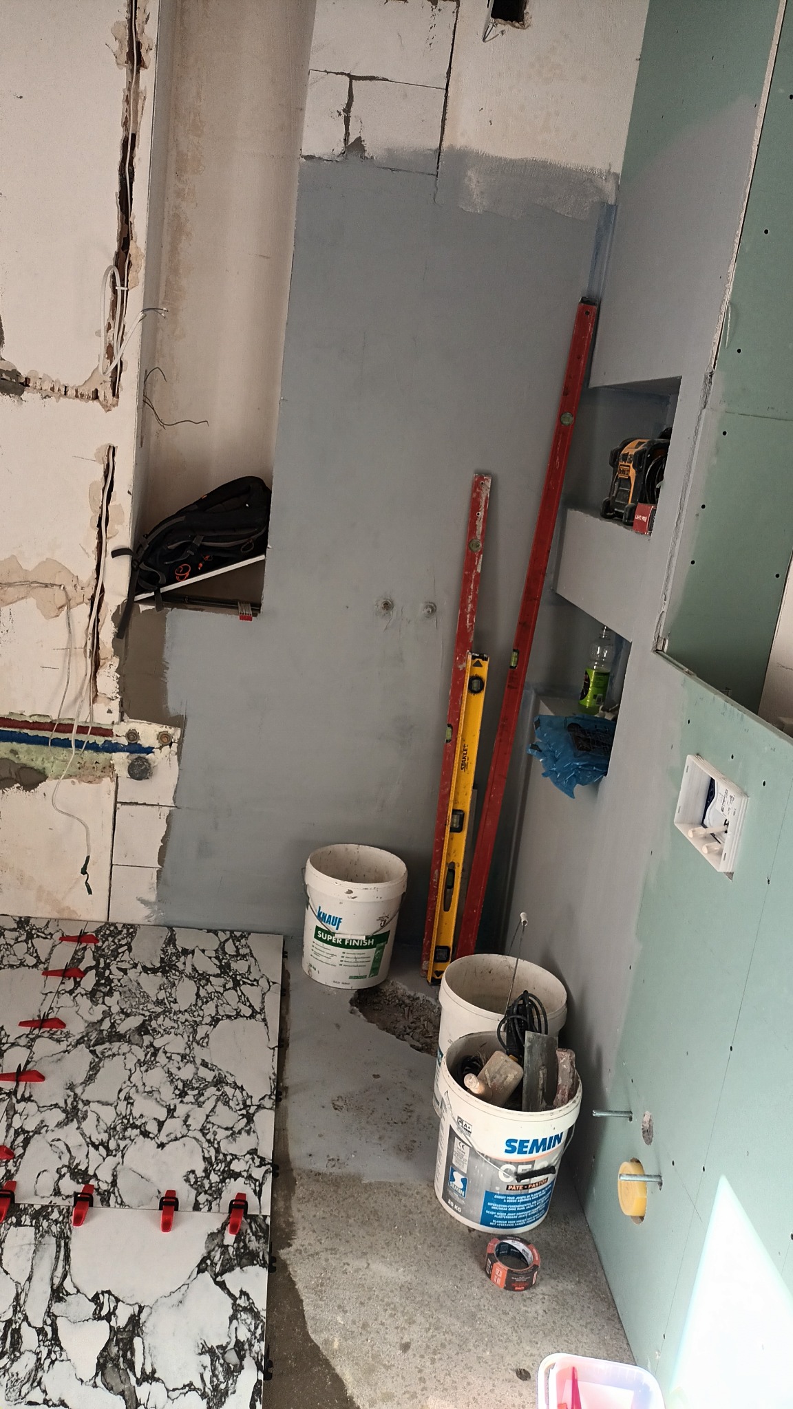 Fragment łazienki w trakcie prac: ułożone płytki imitujące marmur z czerwonymi krzyżykami dystansowymi, ściana częściowo pokryta szarą gładzią, widoczne poziomice, puszki z materiałami budowlanymi...
