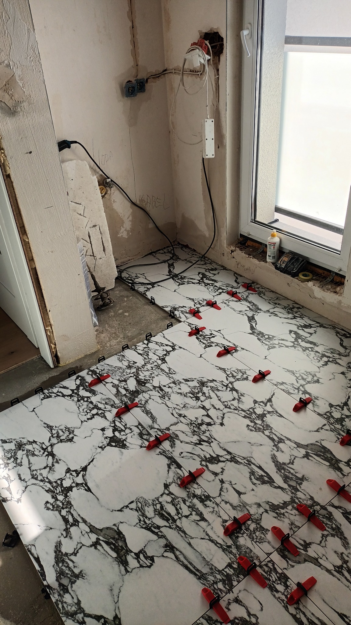 Układanie białych płytek podłogowych imitujących marmur, z czerwonymi krzyżykami dystansowymi, widoczne niedokończone ściany i instalacje elektryczne.