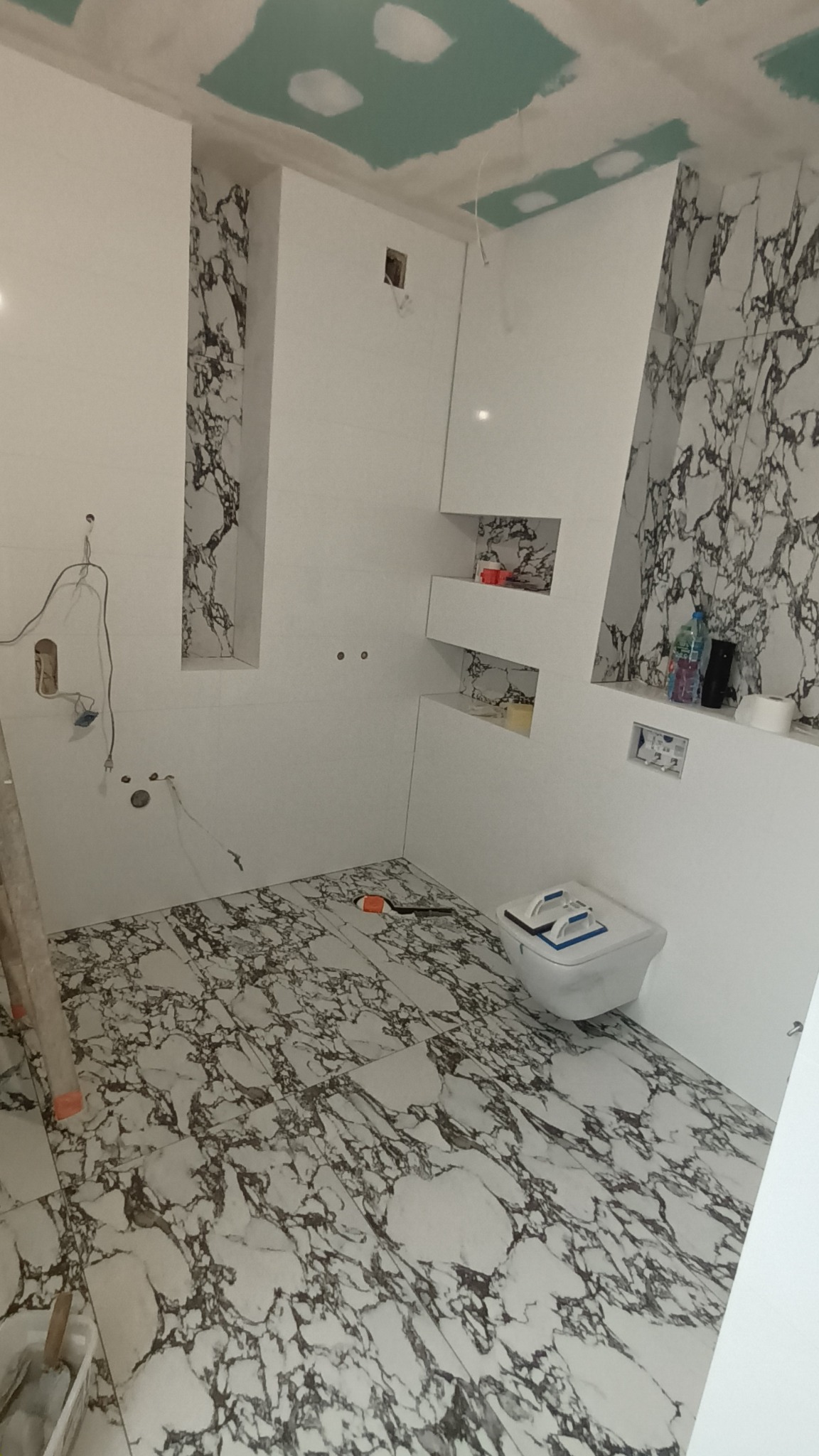 Wnętrze łazienki w trakcie prac wykończeniowych, widoczne płytki imitujące marmur na podłodze i ścianach, niezamaskowane otwory na gniazdka, częściowo wykończony sufit z płyt kartonowo-gipsowych...