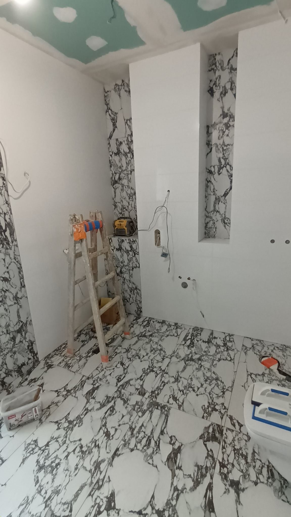 Łazienka w trakcie prac wykończeniowych, widoczne płytki imitujące marmur na podłodze i częściowo na ścianach, białe płytki, drabina oparta o ścianę, narzędzia i materiały budowlane.
