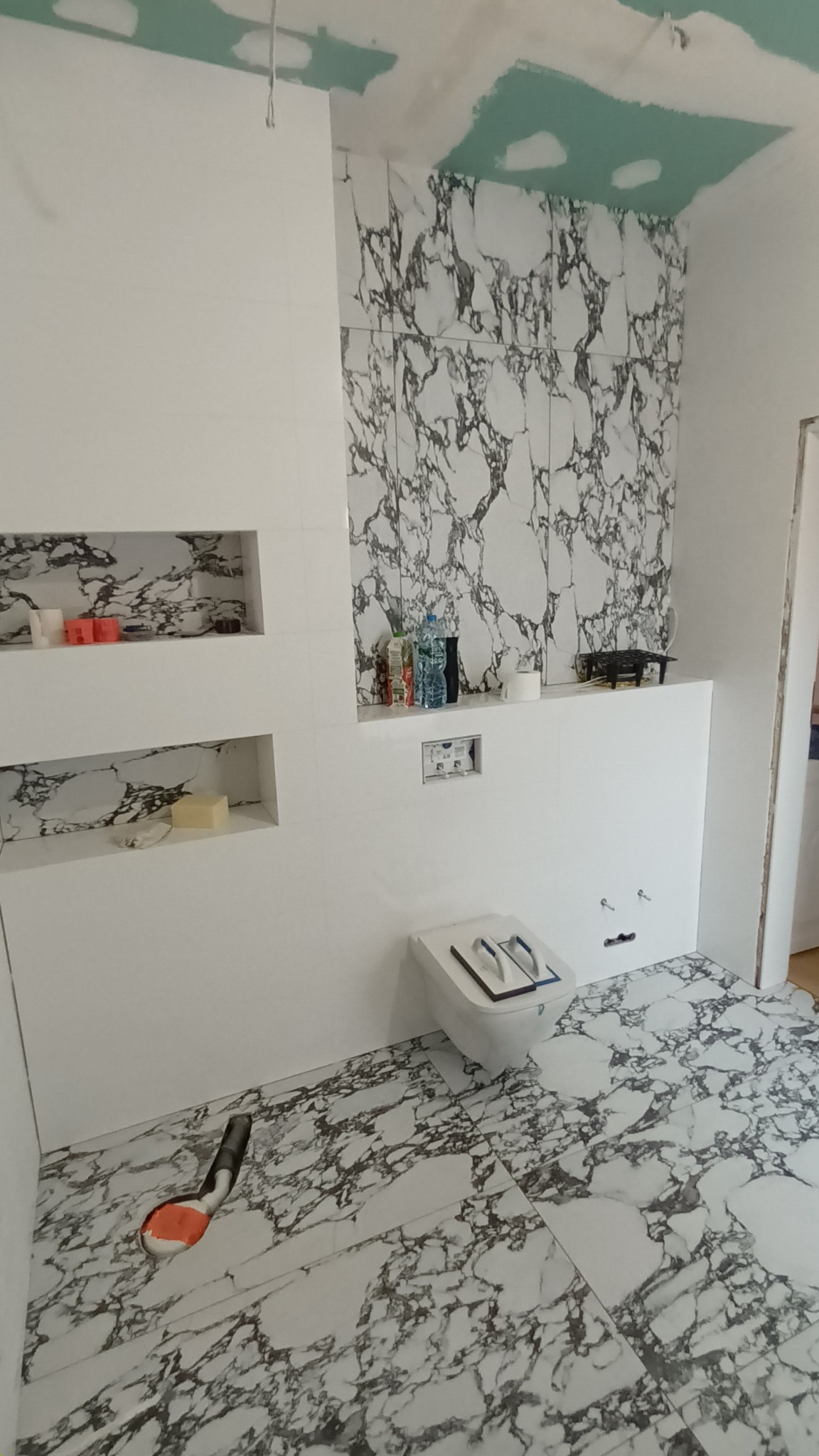 Wnętrze łazienki w trakcie wykańczania z marmurowymi płytkami na podłodze i ścianach, widoczne półki w ścianie, podwieszana toaleta i niedokończony sufit z płyt kartonowo-gipsowych.