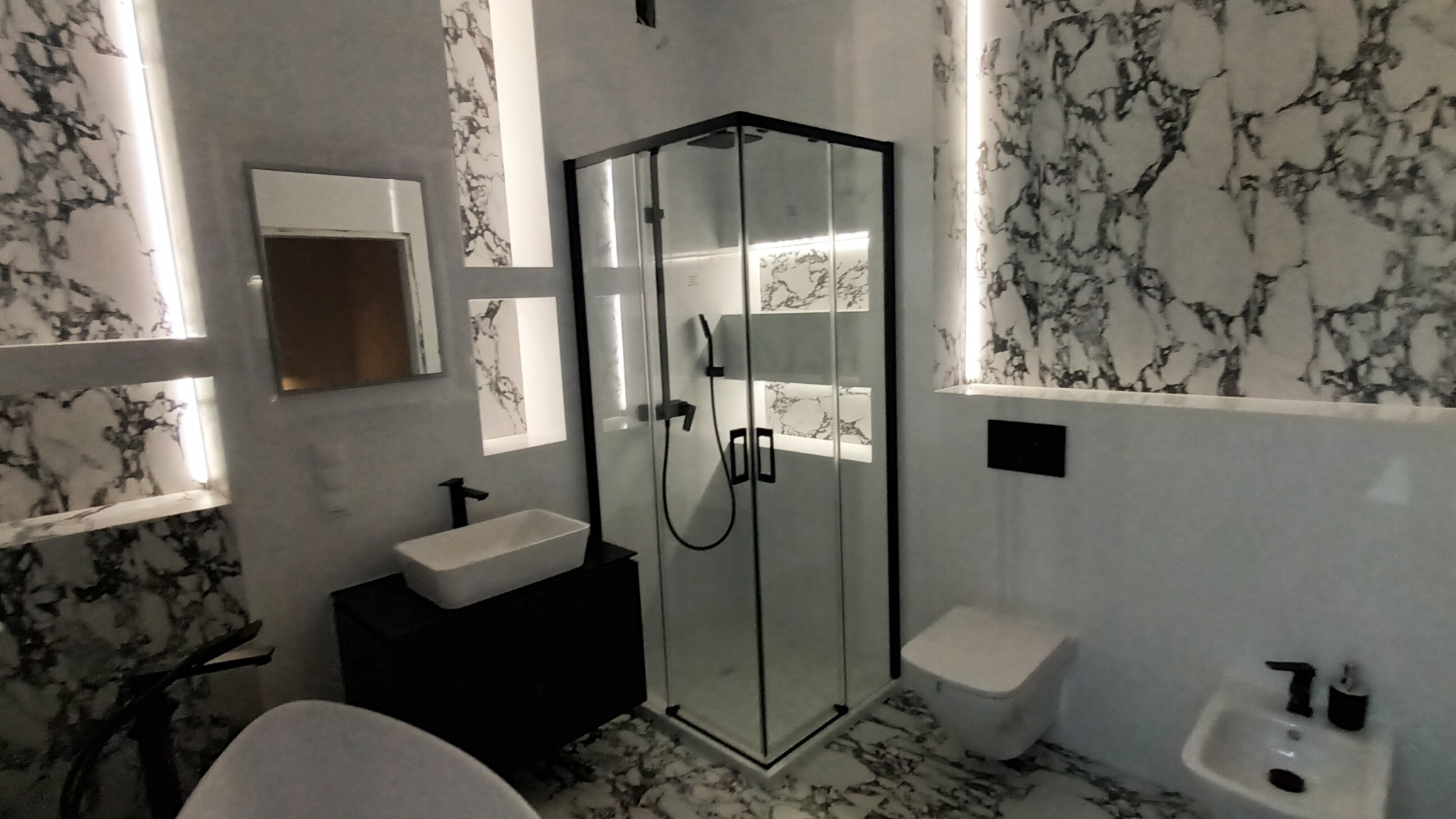 Nowoczesna łazienka w bieli i czerni z marmurowym wzorem na ścianach i podłodze, kabina prysznicowa z czarnymi ramami, umywalka nablatowa, toaleta podwieszana.