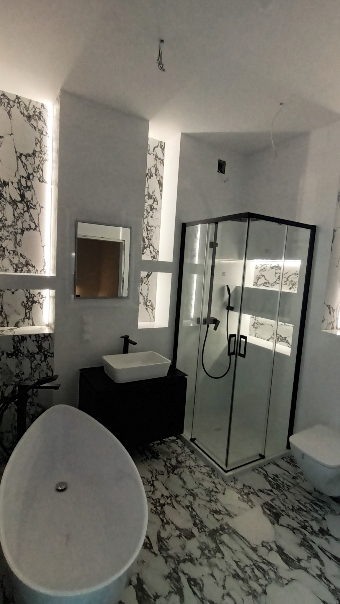 Nowoczesna łazienka z marmurowym wykończeniem, wolnostojąca wanna, kabina prysznicowa z czarnymi profilami, umywalka nablatowa i podświetlane nisze w ścianie.