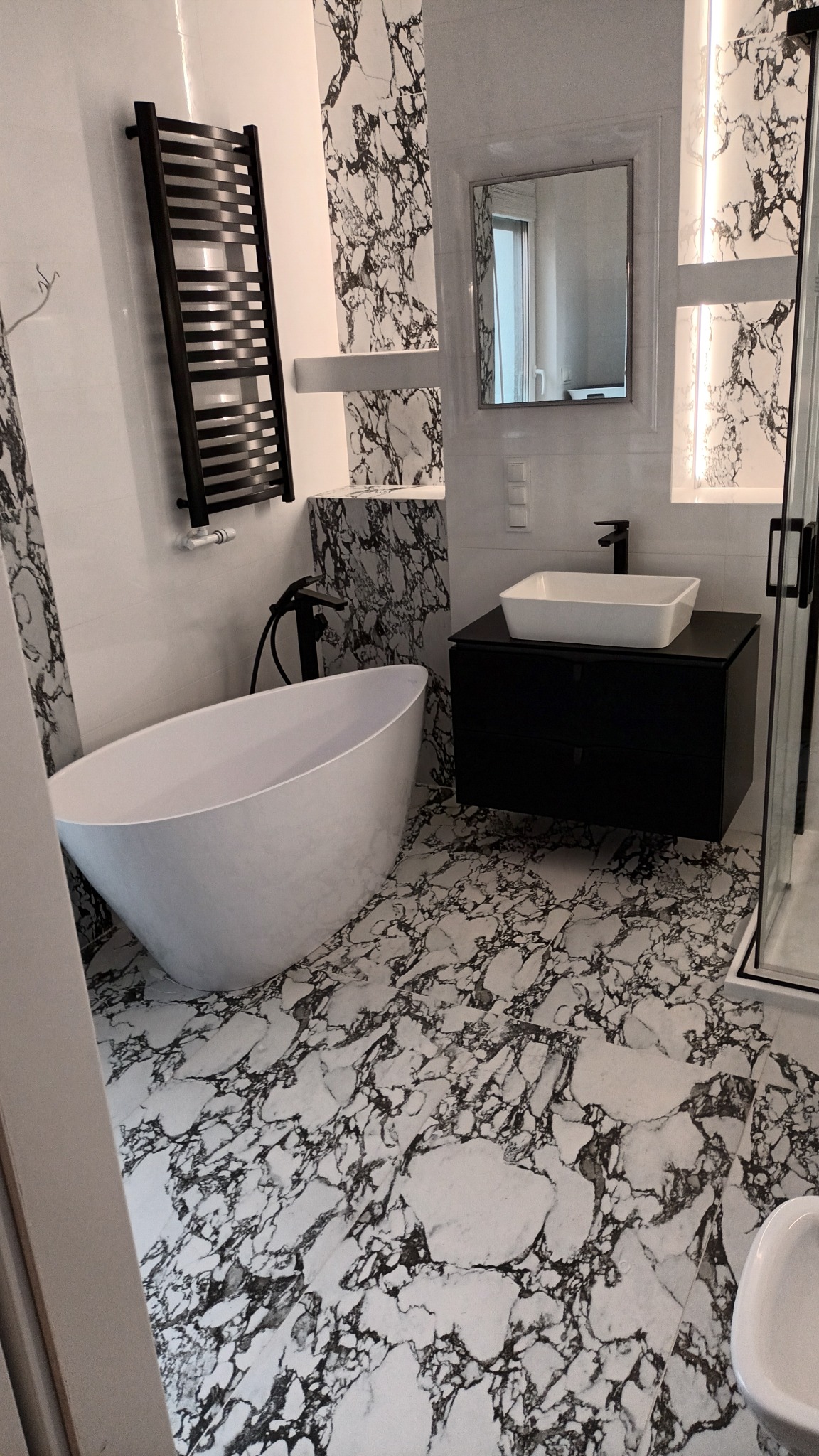 Nowoczesna łazienka z wolnostojącą białą wanną, czarną armaturą i umywalką nablatową na czarnej szafce, wykończona płytkami imitującymi marmur Carrara na podłodze i ścianach.