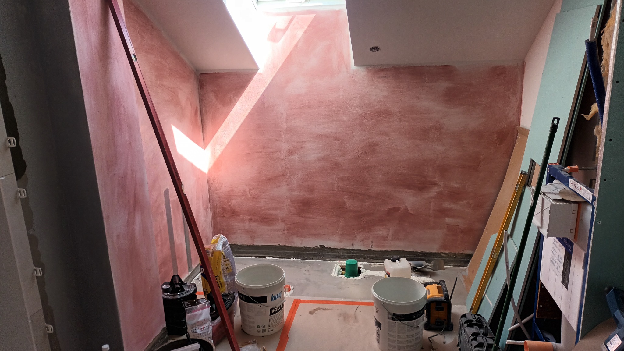 Ściany wstępnie przygotowane pod gładzie w łazience z oknem dachowym. Widoczne wiadra z zaprawą, poziomica i płyty gipsowe.