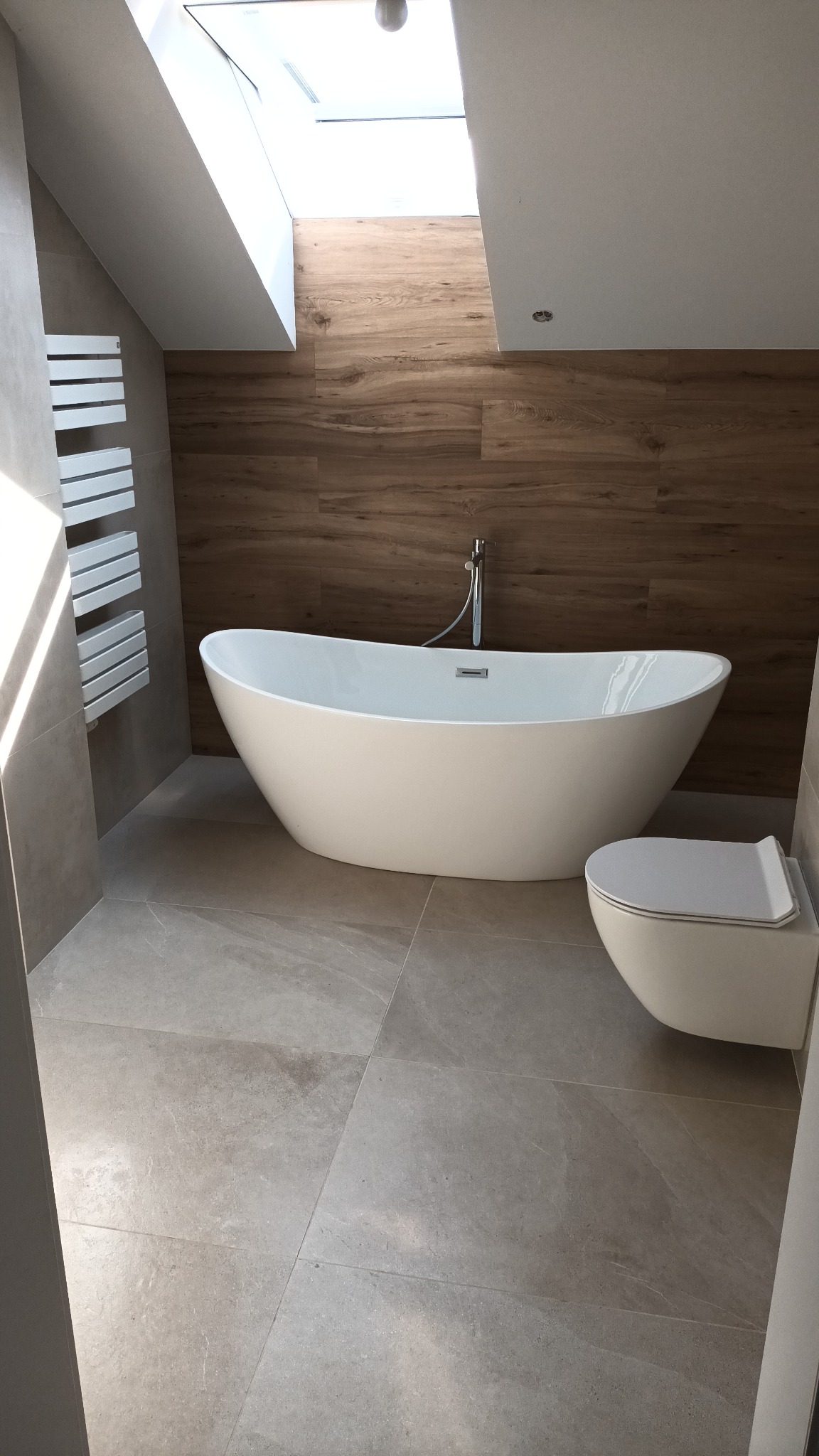 Nowoczesna łazienka w Tychach z wolnostojącą wanną, drewnopodobnymi płytkami na ścianie i grzejnikiem drabinkowym. Jasne płytki podłogowe i okno dachowe.