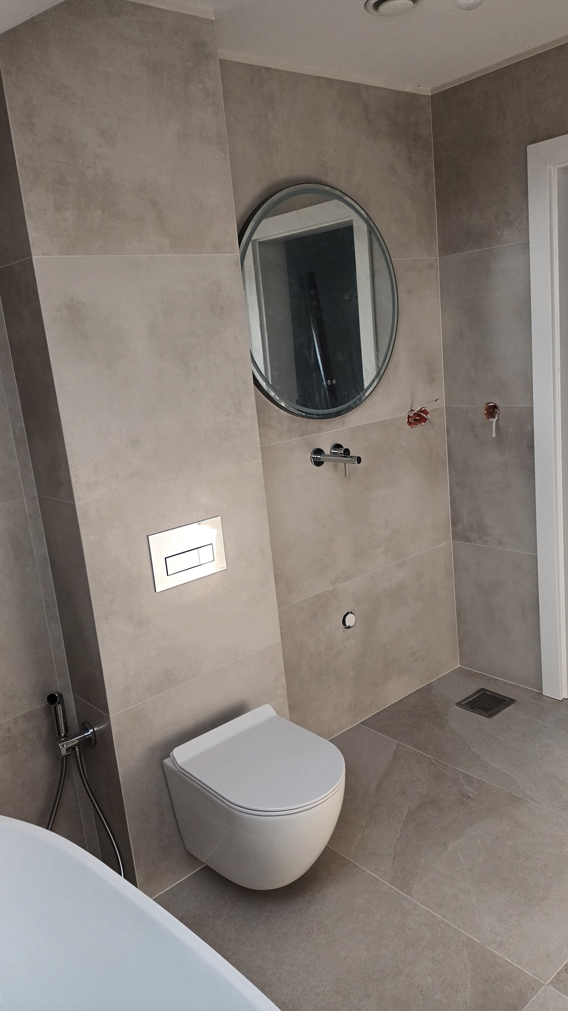 Nowoczesna łazienka z podwieszanym WC, okrągłym lustrem i beżowymi płytkami. Widoczne elementy instalacji wodnej i odpływ podłogowy.