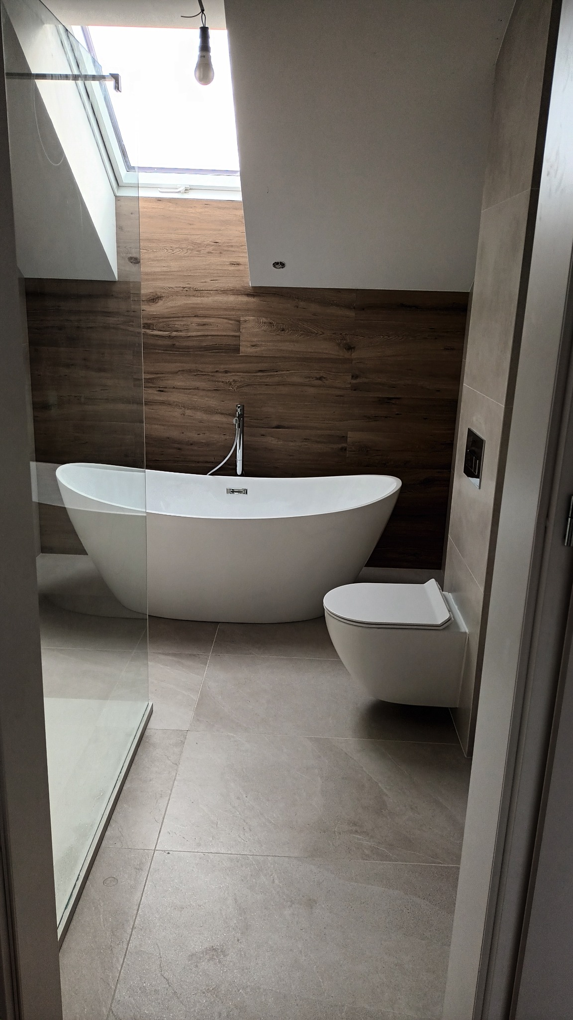 Nowoczesna łazienka z wolnostojącą wanną i toaletą w zabudowie, drewnopodobne płytki na ścianie, szklana kabina prysznicowa, okno dachowe. Stonowana kolorystyka.