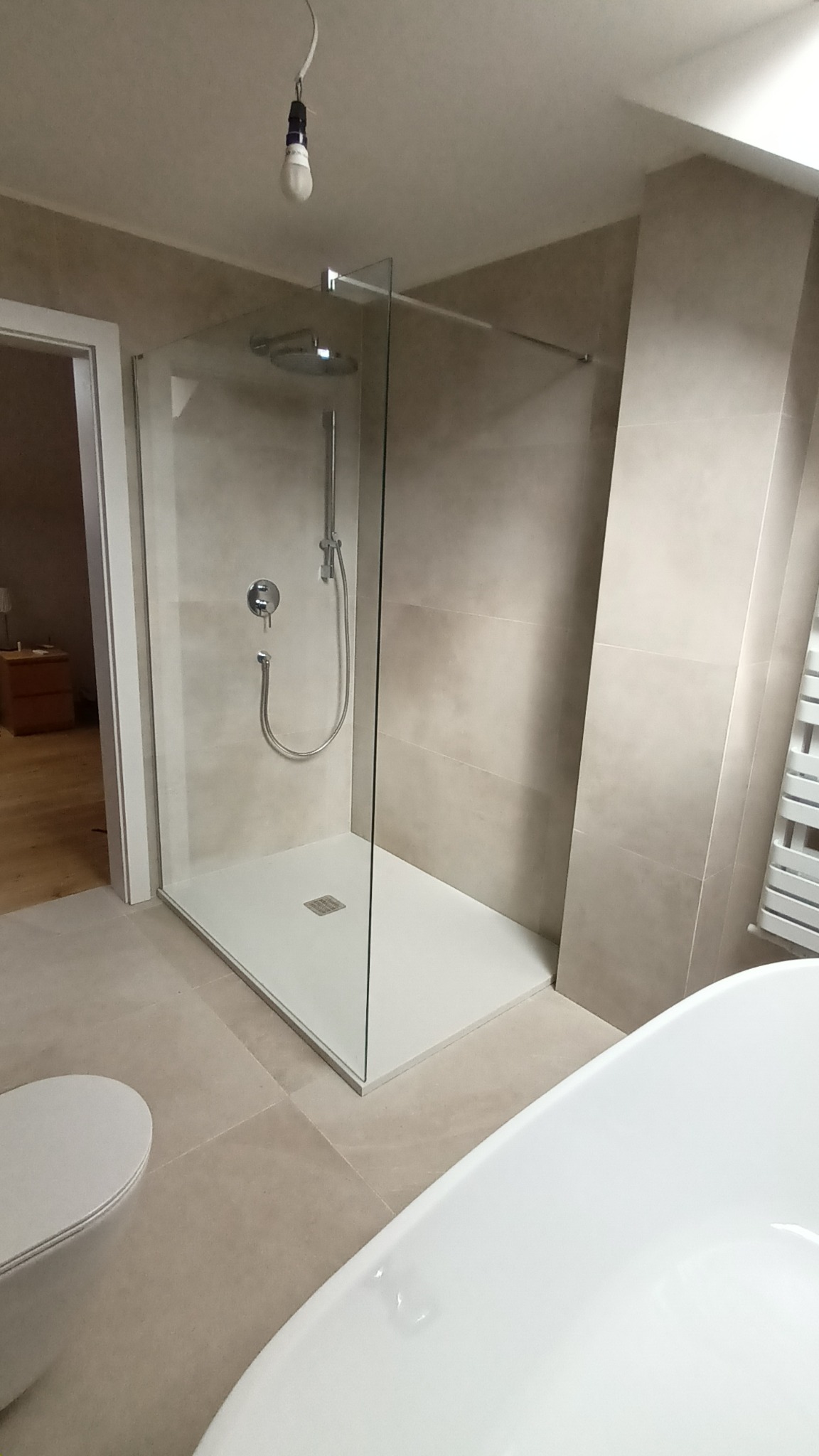 Beżowa łazienka po remoncie: kabina prysznicowa typu walk-in z deszczownicą, biała wanna i toaleta. Widoczna żarówka na kablu. Minimalistyczny design.