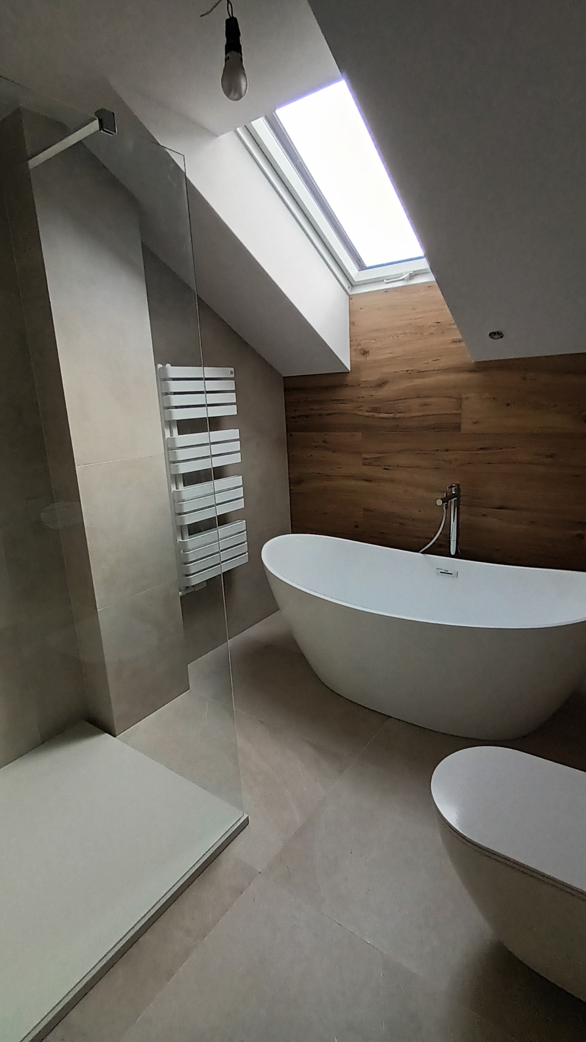 Nowoczesna łazienka na poddaszu z wolnostojącą wanną, kabiną prysznicową i drewnianą okładziną ściany. Widoczne okno dachowe i grzejnik drabinkowy.