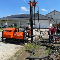 Pomarańczowa wiertnica na gąsienicach podczas pracy na zewnątrz, z widocznymi wężami i pracownikiem w czerwonej koszulce, na tle nowoczesnego domu z panelami słonecznymi.