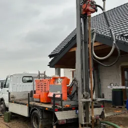 Wiertnica zamontowana na samochodzie ciężarowym podczas wiercenia studni na działce z nowym domem w tle, widoczny błotnisty teren wokół wiertła.