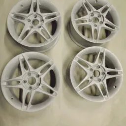 Cztery białe, pięcioramienne felgi aluminiowe po piaskowaniu, leżące na jasnym podłożu.
