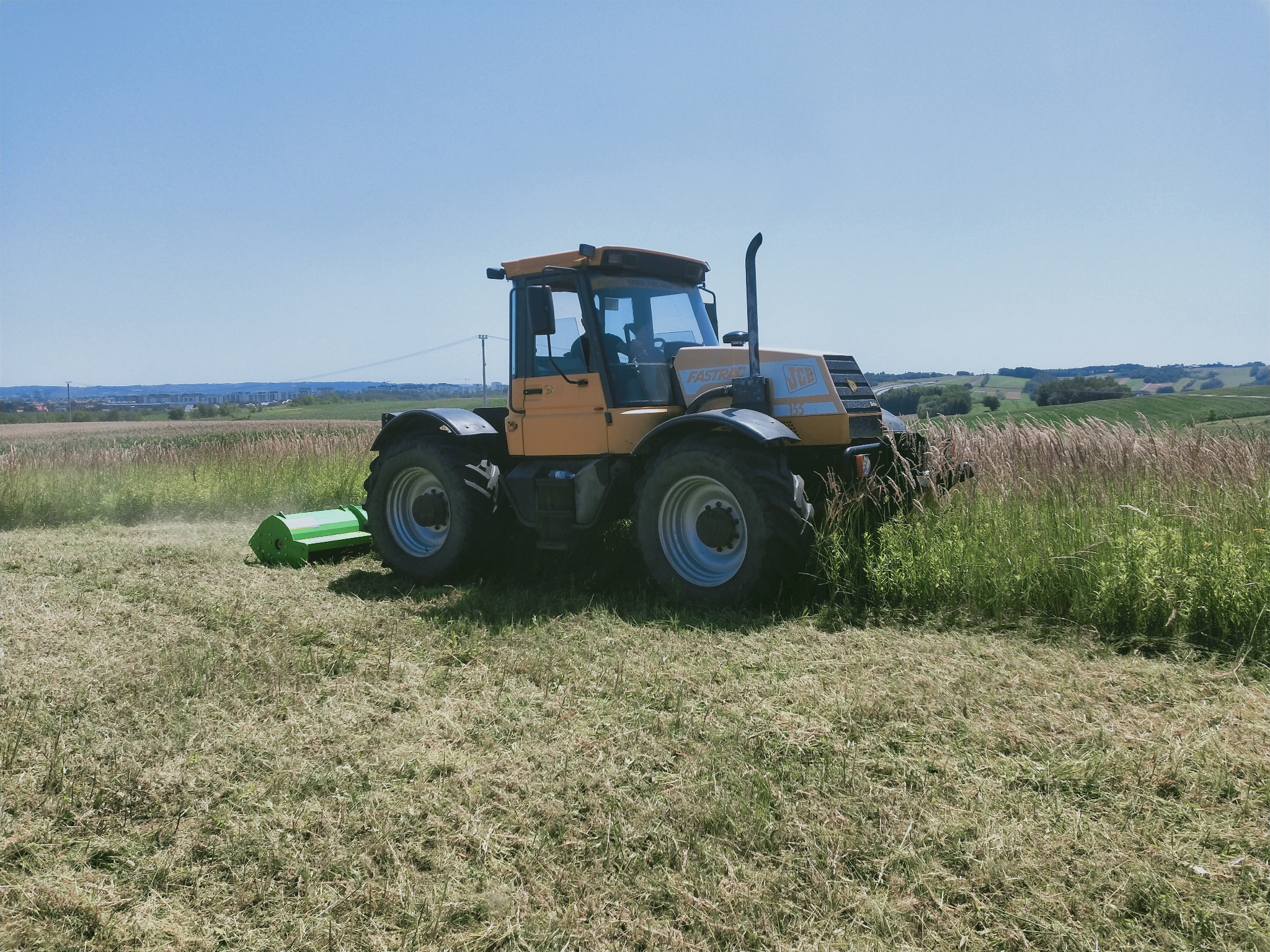 Żółty traktor JCB Fastrac z kosiarką rotacyjną w trakcie koszenia trawy na polu, widoczne ślady świeżo skoszonej trawy, w tle zabudowania miejskie.