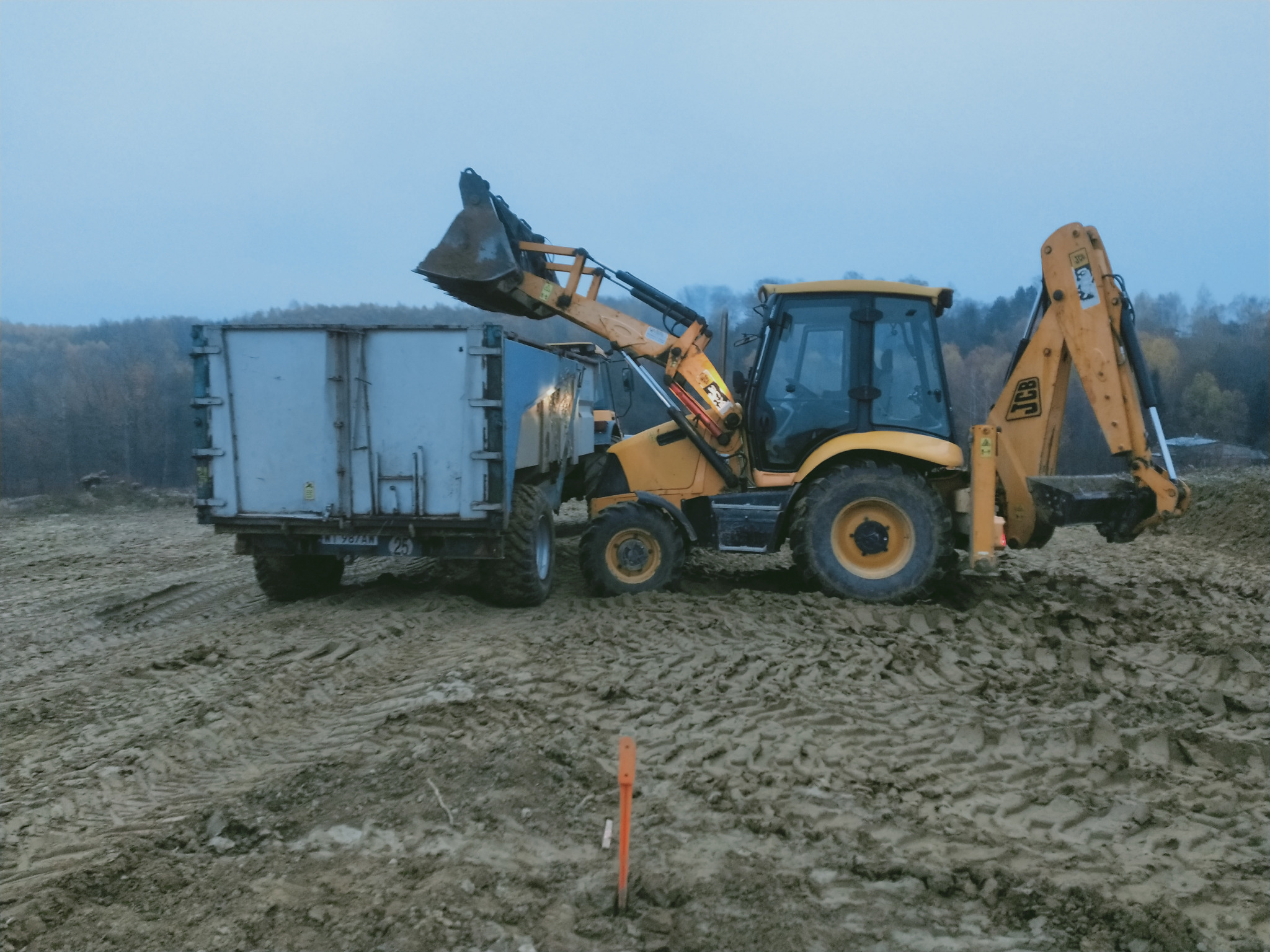 Żółta koparko-ładowarka JCB z podniesioną łyżką i przyczepą na grząskim, błotnistym terenie w pochmurny dzień.