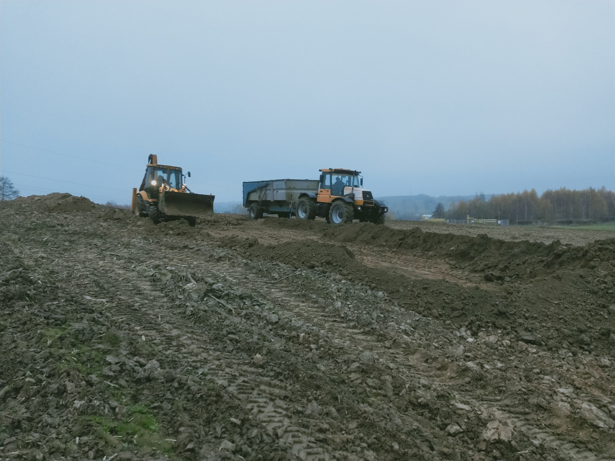 Żółta ładowarka czołowa i ciągnik z przyczepą na polu w trakcie prac ziemnych, widoczne ślady opon na grząskim terenie, pochmurne niebo w tle.