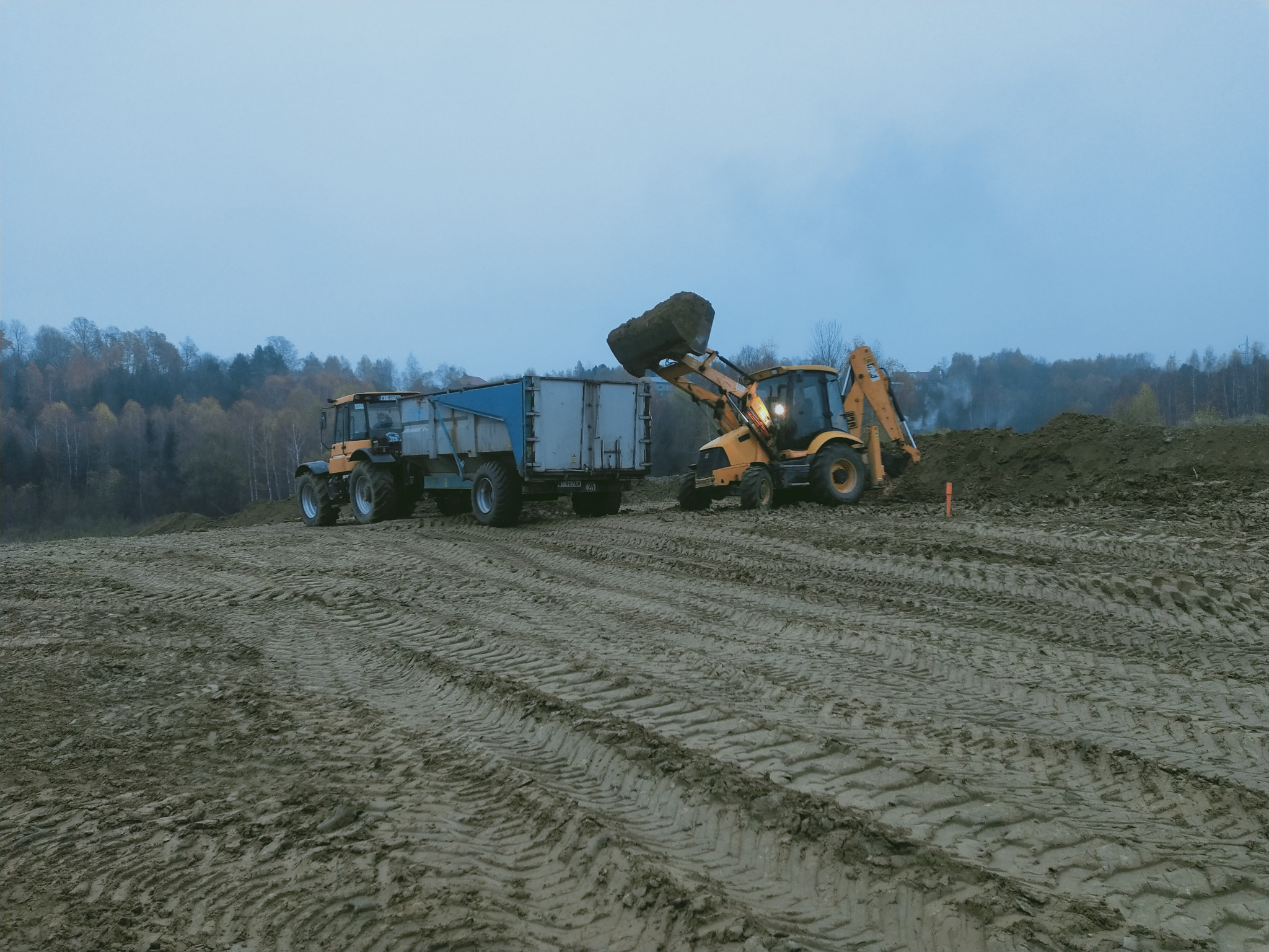 Żółta koparko-ładowarka z podniesioną łyżką pełną ziemi, obok ciągnik z niebieską przyczepą na polu z widocznymi śladami opon, w tle drzewa i pochmurne niebo.