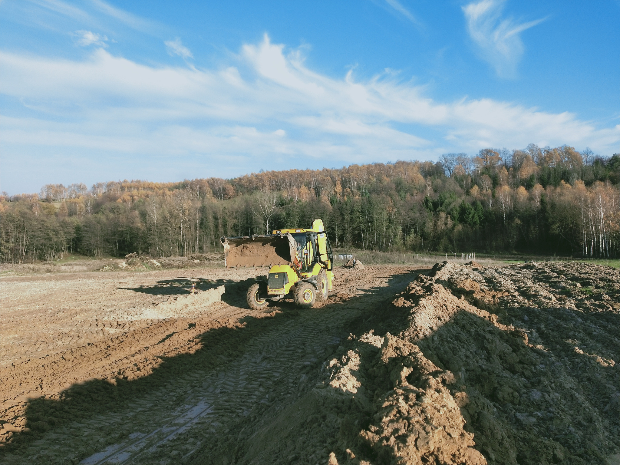 Żółta ładowarka teleskopowa z łyżką pełną ziemi na rozoranym polu, w tle las i błękitne niebo z chmurami.