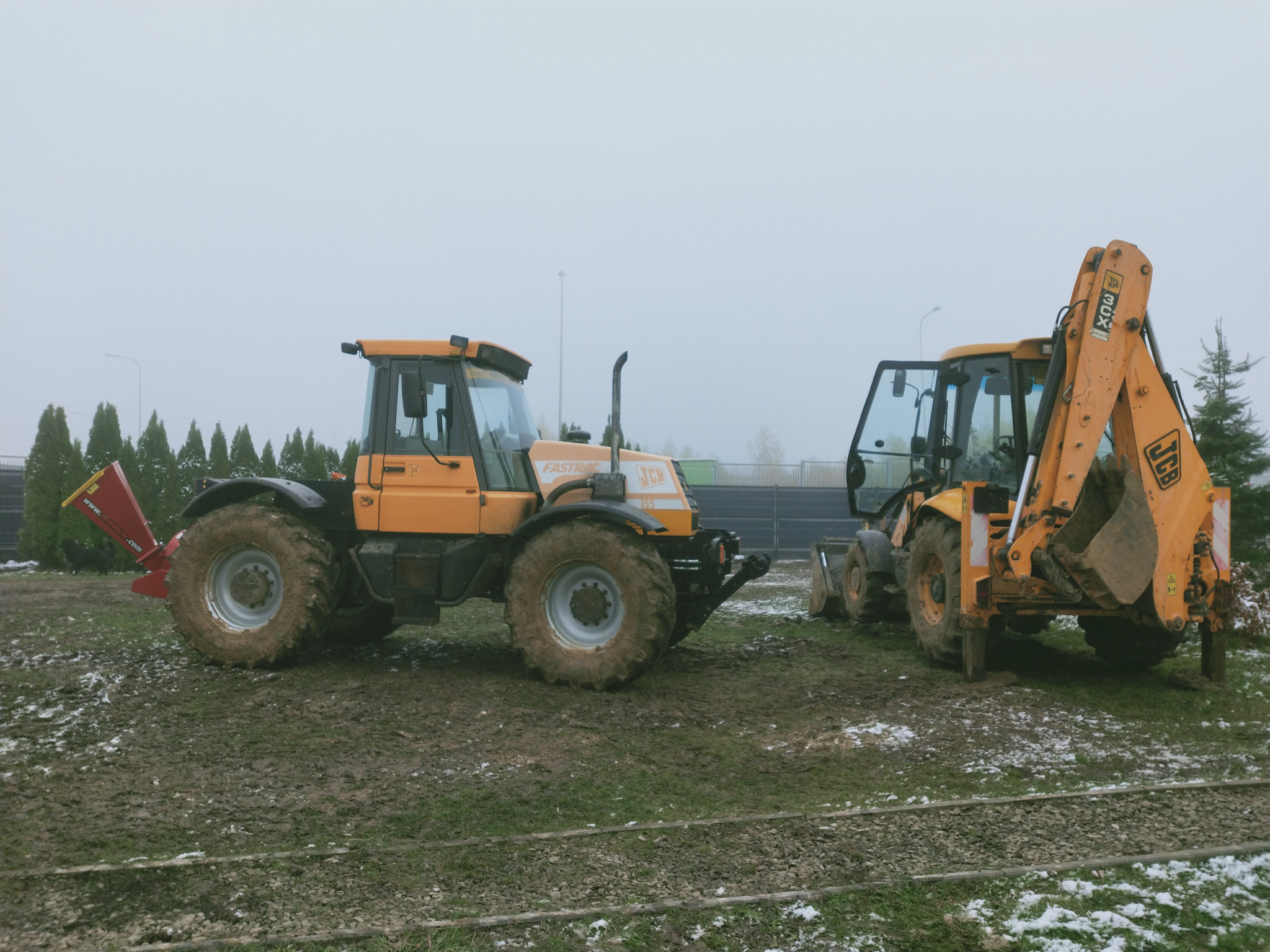 Dwie pomarańczowe maszyny budowlane JCB na błotnistym terenie z resztkami śniegu, jedna z zamontowanym rozdrabniaczem gałęzi, druga z łyżką z przodu i koparką z tyłu, pochmurne niebo w tle.