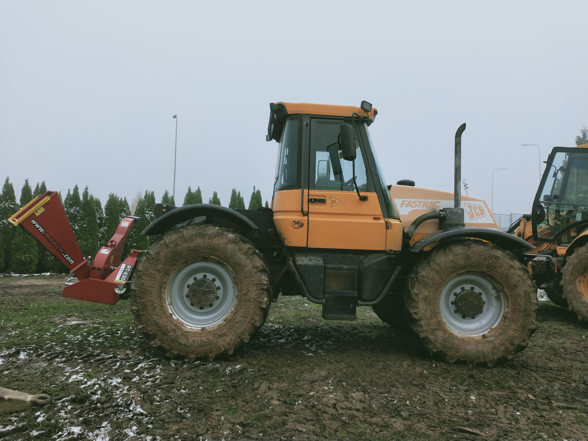 Pomarańczowy ciągnik JCB Fastrac z zamontowanym czerwonym rębakiem do gałęzi na błotnistym terenie, widok z boku w pochmurny dzień.