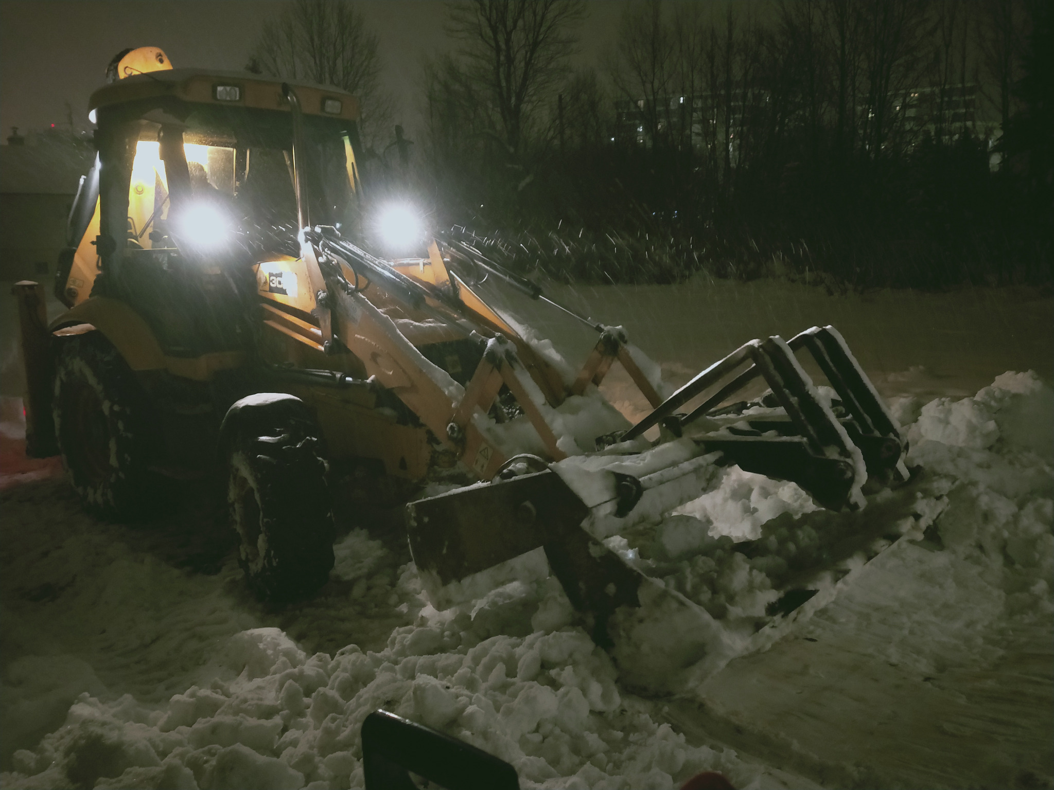 Żółta koparko-ładowarka z włączonymi światłami usuwa śnieg w nocy, widoczne ślady opon i pryzmy śniegu.