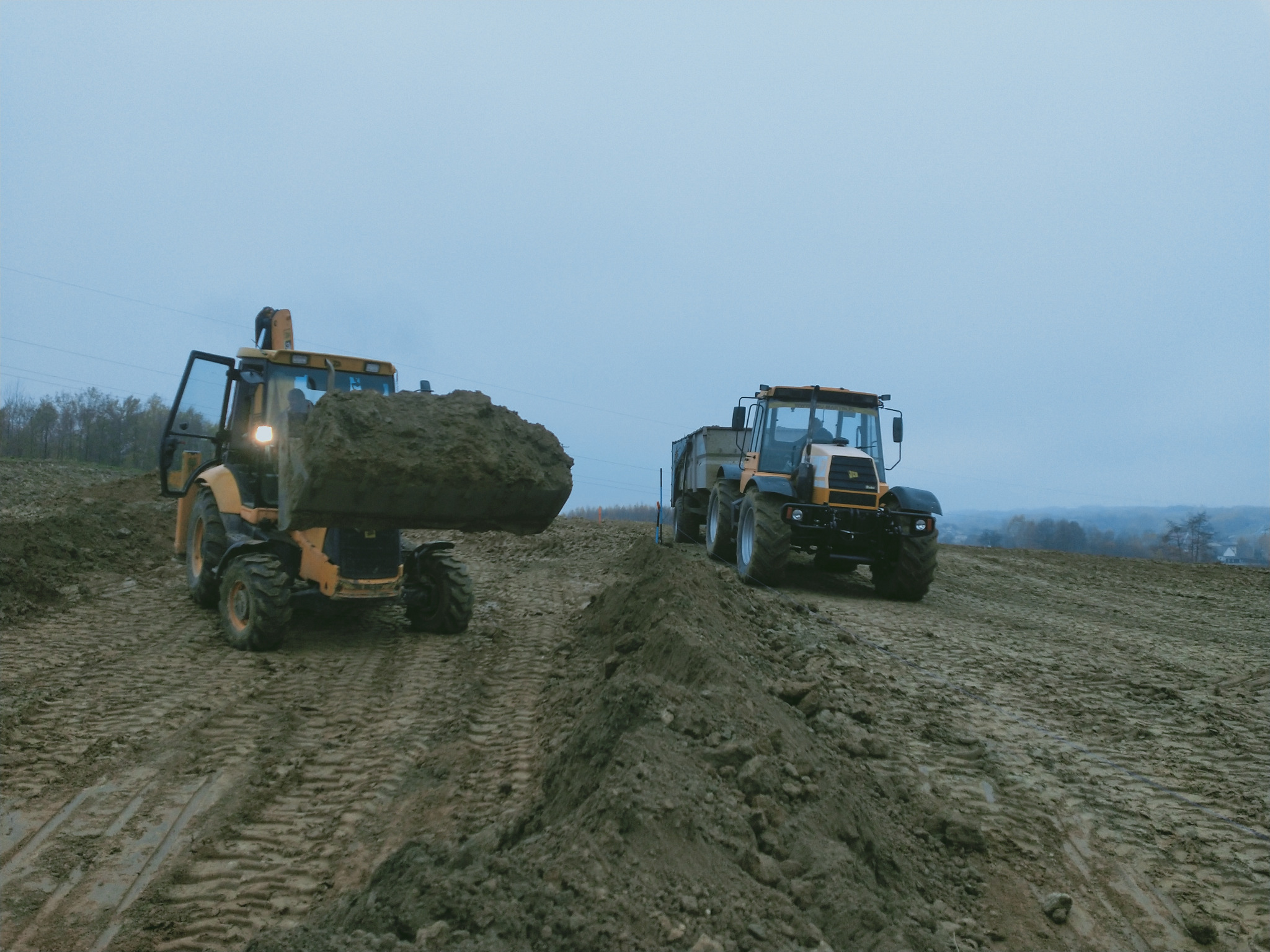 Żółto-czarna koparko-ładowarka z podniesioną łyżką pełną ziemi i czarny traktor z przyczepą na nierównym, błotnistym terenie w pochmurny dzień.