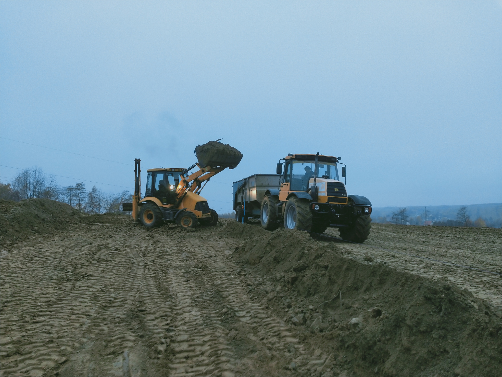 Żółta koparko-ładowarka nabiera ziemię do łyżki, obok stoi traktor z przyczepą, przygotowany do załadunku, w tle widoczne drzewa i zabudowania.