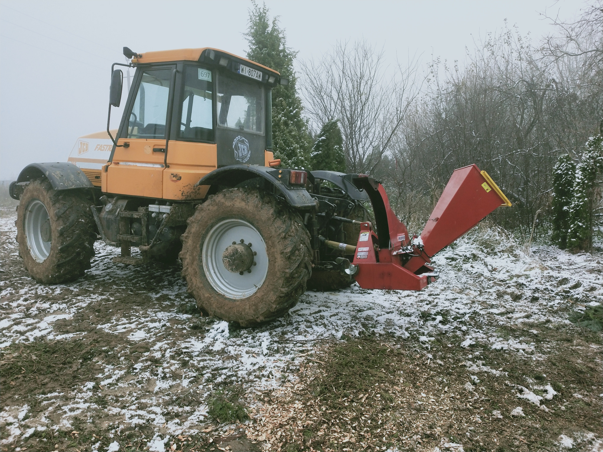 Żółty ciągnik JCB Fastrac z czerwoną rębarką do gałęzi na błotnistym terenie z resztkami śniegu, w tle drzewa i krzewy w pochmurny dzień.