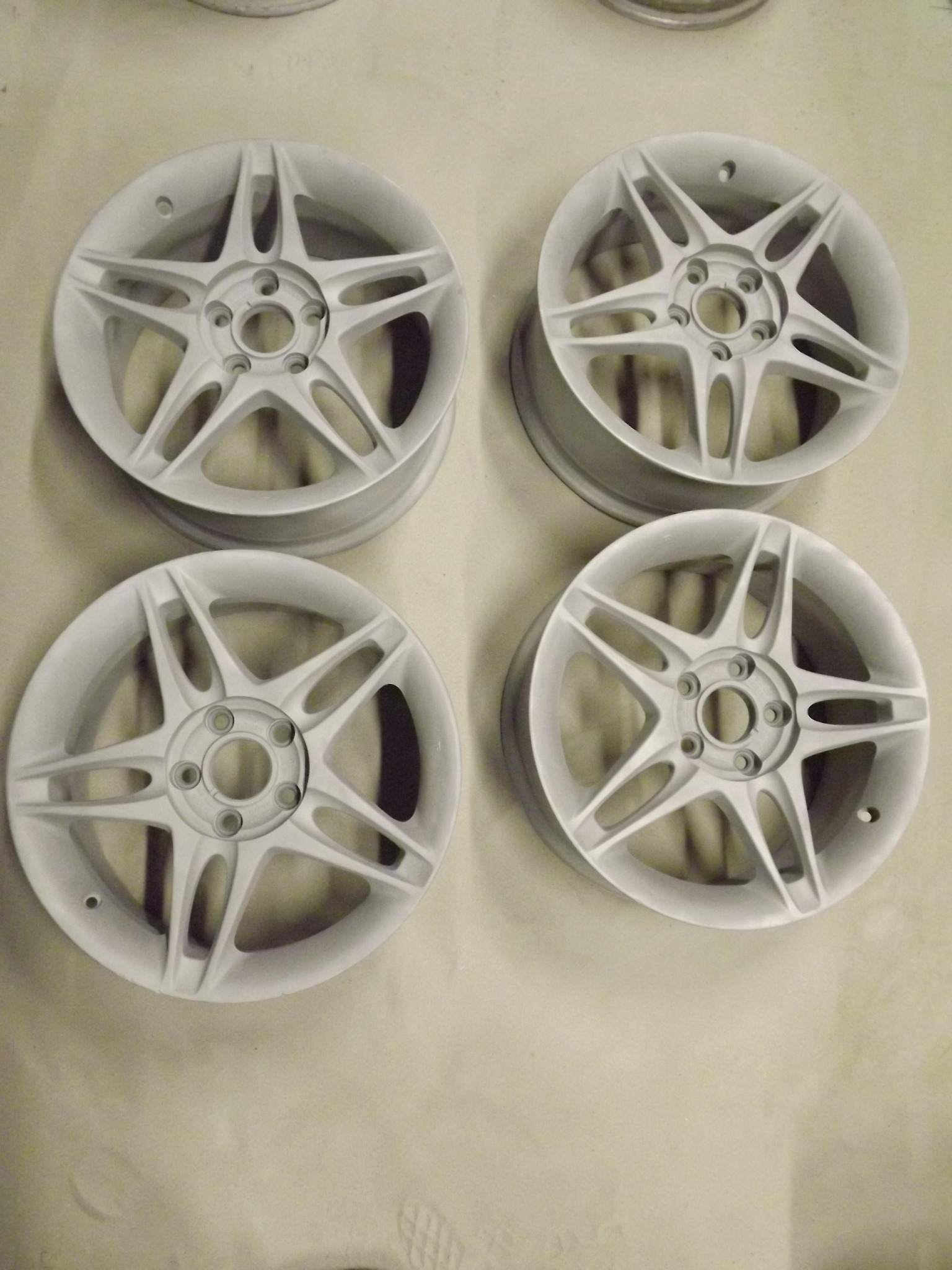 Cztery białe, pięcioramienne felgi aluminiowe po piaskowaniu, leżące na jasnym podłożu.