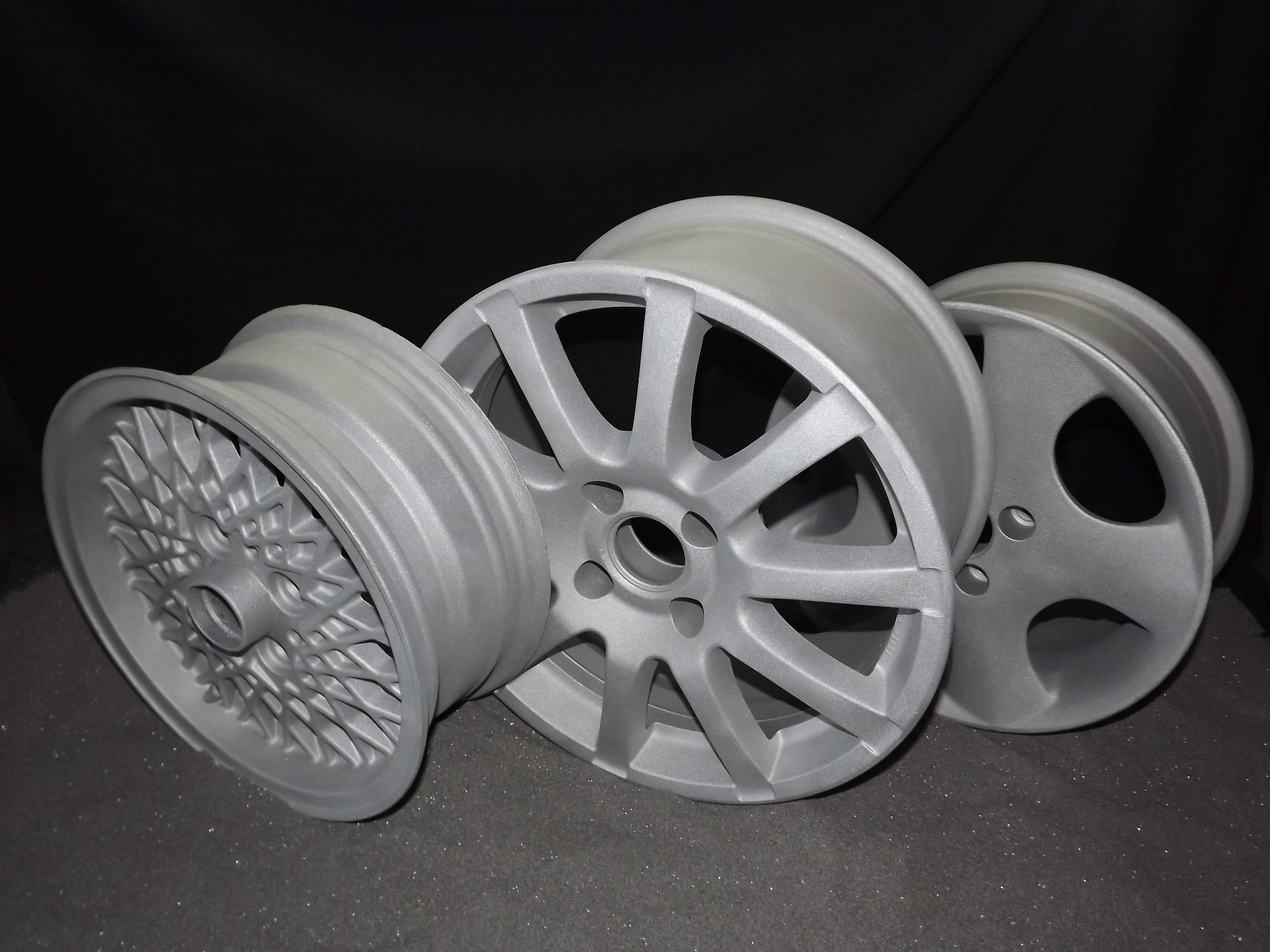 Trzy aluminiowe felgi samochodowe po procesie czyszczenia i nałożeniu podkładu, przygotowane do malowania proszkowego, leżące na szorstkiej powierzchni w ciemnym otoczeniu.