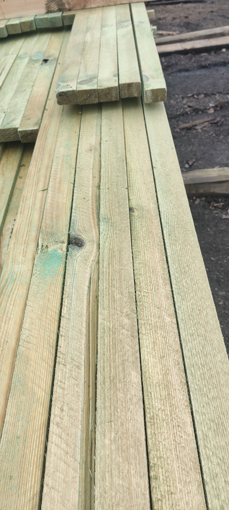 Stos impregnowanej, jasnej tarcicy budowlanej ułożonej na zewnątrz, widoczne sęki i zielonkawy odcień impregnatu.
